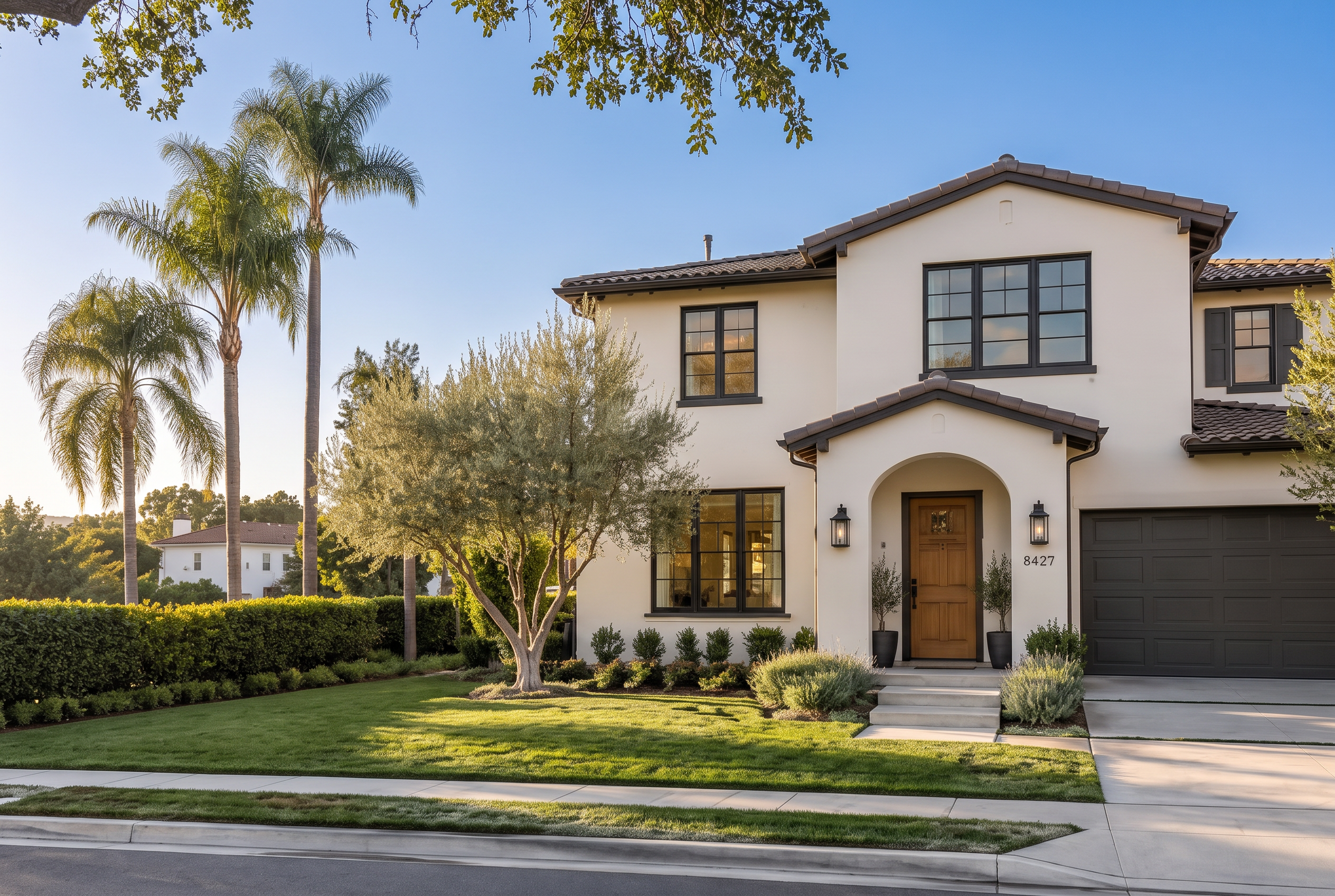 Two-story white stucco home with dark window frames, tile roof and palm trees in warm late-afternoon sun